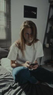 Yatakta cep telefonuyla mesajlaşan, siber zorbalık yüzünden gergin ve sinirli genç kızın dikey görüntüsü.