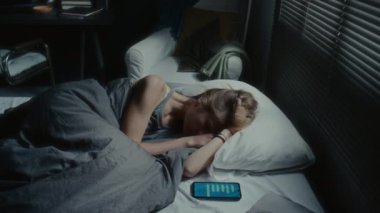 Yüksek açılı bir çekimde, depresif bir kız, tehdit mesajları aldıktan sonra, akıllı telefonuyla yatakta yatar.
