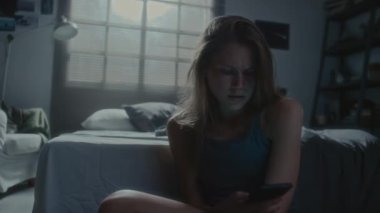 Karanlık bir odada oturmuş, küfürlü mesajlar okuyan, telefonu kaldıran ve siber zorbalık görürken dizlerine sarılan üzgün genç kızın el kamerasıyla çekilmiş fotoğrafı.