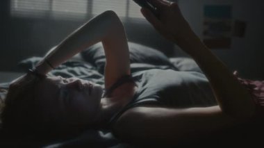 Genç bir kadın yatağa uzanıyor, telefonda aşağılayıcı mesajlar okuyor, uykusuz gecelerde üzülüyor ve yan yatıyor.