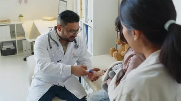 用无接触式红外温度计测量手拿着毛绒玩具坐在医疗沙发上的拉美裔小女孩体温的中东男性儿科医生中值 — 图库视频影像