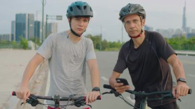 Spor giyim ve koruyucu kasklı baba ve genç çocuğun portresi yolda bisiklete binerken kameraya poz verirken yolda duruyor.