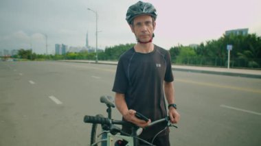 Güvenlik kaskı ve spor giyimli erkek bisikletçinin yolda dikilip, elinde telefonla kameraya poz verirken orta uzunlukta bir görüntüsü.