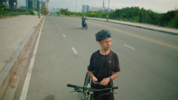 スマートフォンでナビゲーションアプリを使用して 道路上の自転車で立っているスポーツウェアとヘルメットでサイクリストのショットを傾け 周りを見渡す — ストック動画