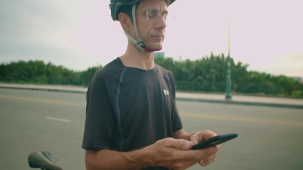 男子自行车手戴着防护头盔和运动服站在室外 在骑自行车休息时用智能手机导航的中景照片 — 图库视频影像