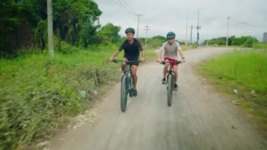 Baba ve genç oğul kask ve spor kıyafetleriyle tropik kırsal bölgelerde bisiklet sürerken yoldan çıkıyorlar.
