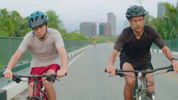 父亲和儿子都戴着防护头盔 骑着运动服 骑着自行车在柏油路上对着摄像机 然后在室外骑车 — 图库视频影像