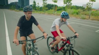 Ön manzara, kasklı bir adam ve çocuğun şehir yolundaki kameraya doğru bisiklet sürüşünü gösteriyor.