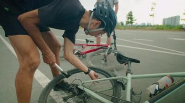 Bisiklet kasklı bir adam bisiklet lastiğini pompayla şişiriyor. Oğlu arka planda su içerken yolda duruyor.