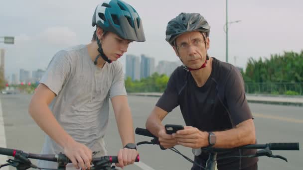 爸爸和儿子穿着运动服 戴着防护头盔 骑着自行车站在路上 在智能手机上使用在线地图 一边选择路线一边环顾四周 — 图库视频影像