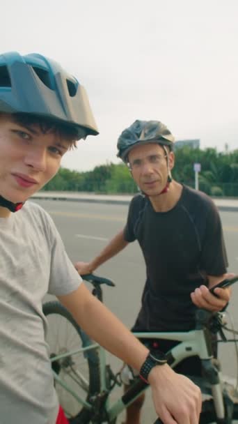 男孩和他的爸爸骑自行车 戴着头盔 穿着衣服 看着相机 一边在拍摄骑自行车旅行的视频一边聊天 一边垂直拍摄的Pov照片 — 图库视频影像