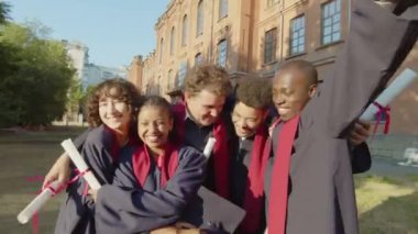 Üniversite mezuniyetini kutlarken birbirlerine sarılıp dışarıda kameralara bakan neşeli arkadaşların portresi.