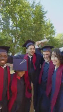 Mezuniyetlerini kutlamak için elbise ve üniversite şapkası takan çok ırklı mutlu arkadaşların dikey çekimi.