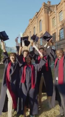 Mutlu mezun öğrencilerin dikey portresi şapkalarını ve diplomalarını kaldırıyorlar ve tören üstüne kamera izliyorlar.