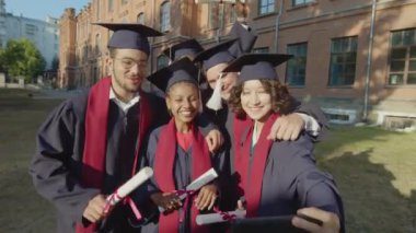 Açık havada akıllı telefondan selfie çekmek için bir araya gelen çok ırklı mezun öğrencilerin görüntülerini yakınlaştırın.