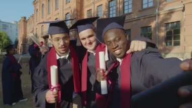 Çok ırklı erkek arkadaşların cep telefonuyla selfie çekerken, mezuniyet süresince üniversitenin önünde dikilirken orta boy bir fotoğraf.