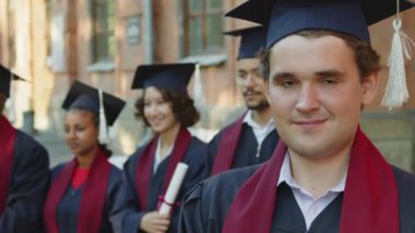 Kafkasyalı erkek öğrencinin portresi, kolej arkadaşlarının yanında duruyor, önlüklü ve şapkalı ve kameraya bakıyor.