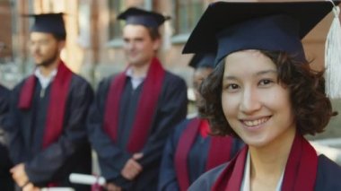 Şapkalı ve cüppeli neşeli bir bayanın yakın portresi kameraya bakıyor, diğer öğrencilerin yanında durup veda konuşmasını dinliyor.