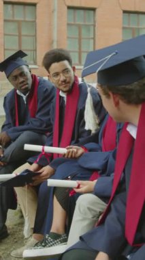 Mezuniyet töreninden sonra kampüste otururken iletişim kuran neşeli öğrencilerin dikey çekimi.