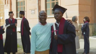 Babası ile birlikte üniversiteye karşı fotoğraf çektiren önlüklü ve şapkalı Afrikalı genç bir adamın portresi.