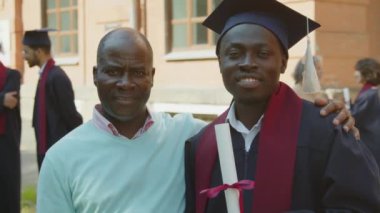 Orta ölçekli, neşeli siyahi mezun öğrencinin üniversitede babasıyla fotoğraf çektirmek için poz vermesi.