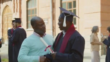 Orta yaşlı bir babanın mezuniyetinde genç oğlunu cübbe ve üniversite şapkasıyla tebrik ettiği orta boy bir fotoğraf.