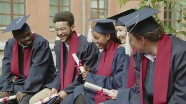 Üniversiteye yakın bir yerde tören beklerken gelecek planlarını tartışan çok kültürlü genç öğrencilerin orta dereceli fotoğrafı.