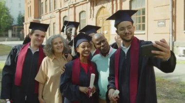 Orta boy bekar bir erkeğin üniversite arkadaşları ve aileleriyle dışarıda akıllı telefon kullanarak selfie çektiği bir fotoğraf.