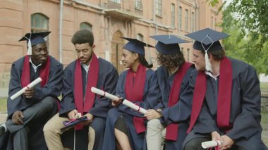 Kampüste mezuniyet törenini beklerken iletişim kuran cüppeli ve şapkalı beş çok kültürlü öğrencinin orta ölçekli görüntüsü.