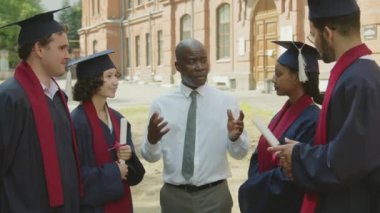 Orta boy Afro-Amerikan profesörün üniversitenin dışındaki tören sonrası elbise ve şapka giyen öğrencilerle iletişim kurduğu bir fotoğraf.