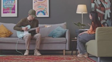 Oturma odasındaki kanepede gitar çalan ve evde özel ders sırasında bayan müzik öğretmeninin talimatlarını dinleyen genç bir adam.