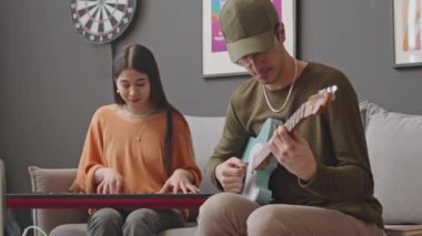 Gitar çalan genç adamın görüntüsünü yakınlaştır ve kadın müzisyen ile ev stüdyosunda synthesizer çalarken konuş.