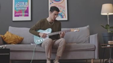 Elektro gitar çalan ve oturma odasındaki kanepede otururken kameraya poz veren genç adamın portre fotoğrafını yakınlaştır.