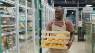 Orta boy Afro-Amerikan erkek süpermarket çalışanı taze olgun portakal dolu tahta bir sandık taşırken buzdolaplarıyla koridorda yürürken, meyve ve sebzelerin sabah planlarını yaparken.