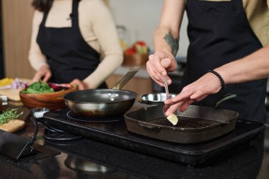 Tanımlanamayan bir adamın, arka planda sebze kesen bir kadınla mutfak sobasının yanında duran bir parça tereyağı koyması.