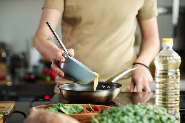 Closeup of unrecognizable man pouring egg liquid from bowl into frying pan while standing by stove at kitchen