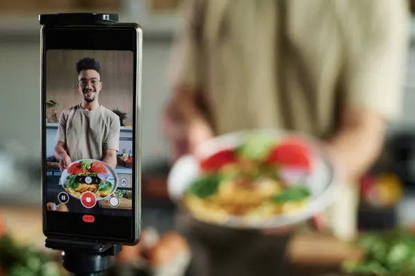 Closeup of mobile phone on tripod filming young middle eastern blogger showing plate with omelet and vegetables, focus on smartphone, copy space