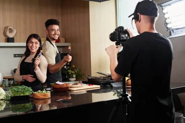 Middle eastern man and hispanic woman standing back to back holding forks with food by kitchen counter while being photographed by cameraman