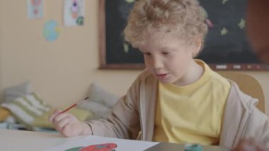 Kıvırcık saçlı sarışının, ilkokulda sanat dersi sırasında bir grup çocukla birlikte masa başında otururken uzay roketi çizdiği pozu.