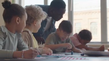 İlkokulda resim çizerken eğlenen neşeli çoklu etnik çocuklar, geride kalan Afrikalı Amerikalı öğretmenler,