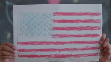 Küçük Afrikalı Amerikalı kız elinde Amerikan bayrağını kameranın önünde çiziyor, sonra yüzünü gösteriyor ve sınıfta gülümseyerek poz veriyor.