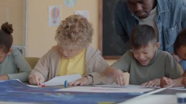 İlköğretim çağında çok ırklı erkek ve kızlar resim dersinde Afrika kökenli Amerikan öğretmeninin kontrol ettiği pastel boyalarla masa başında oturuyorlar.