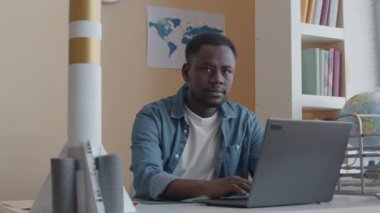 Genç Afrikalı Amerikalı okul öğretmeninin portresi, el yapımı uzay roketi ile masada oturuyor, dizüstü bilgisayarında daktilo çekiyor ve iş gününde kameraya gülümseyerek poz veriyor.