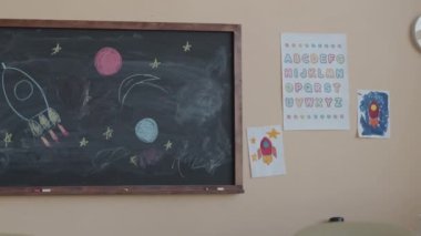 İlköğretim sınıfının içinde duvarda çocuk resimleri ve karatahtaya uzay roketi çizimleri var.
