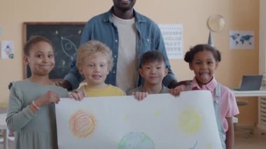 Grup portresi mutlu Afrikalı Amerikalı öğretmen ve çok ırklı çocukların ellerinde gezegenlerin kağıt çizimleri ve sınıfta gülümseyerek kameraya poz vermelerini gösteriyor.