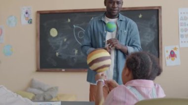 Siyah erkek öğretmen Dünya modelini tutuyor, astronomi dersi sırasında gülümsüyor ve ilkokul öğrencileriyle konuşuyor.