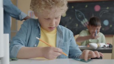 Sarı kıvırcık saçlı küçük çocuk, ilkokulda el işi ödevi yaparken kâğıt çarşafa kurşun kalemle işaret koyuyor.