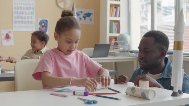 在小学上课期间 非裔美国男教师帮助小女孩在造纸火箭的同时翻纸 — 图库视频影像