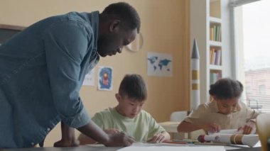 Afrika kökenli Amerikalı öğretmen ilkokulda el işi yaparken küçük Asyalı çocuğa makasla kağıt kesmede yardım ediyor.