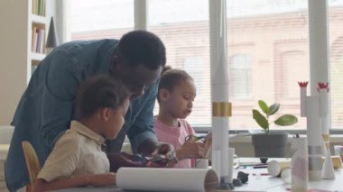 Afrika kökenli Amerikalı öğretmen, sanat ve zanaat dersleri sırasında kağıt roketi yapan iki küçük kıza yardım ederken makasla kağıt kesiyor.